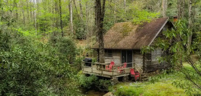 A rustic log cabin is nestled in a forest. The windows and doors are modern, but the wood is aged and grey. A deck is built on one side. Two orange chairs are on the deck. A third orange chair is on the ground next to the stairs leading up to the deck. A tree grows up through the deck and a shelf or table seems to have been constructed around the trunk.